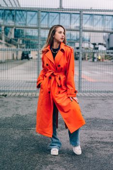 Fashionable blonde woman wearing orange coat, blue jeanse , white sneackers and neckchain posing in street of European city.