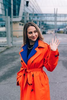 Fashionable blonde woman wearing orange coat, blue jeanse , white sneackers and neckchain posing in street of European city.