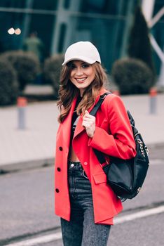 glamour woman in trendy outfit posing against the building urban background, fashion look. Outdoor fashion portrait of stylish young woman wearing black jeans, red jacket, top and a cap