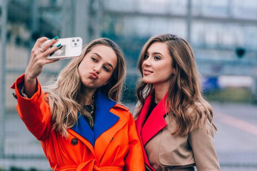 Fashionable happy smiling blonde woman wearing orange coat, blue jeanse and neckchain communicate with her girlfriend on the street. blonde and brunette happy and posing on the street