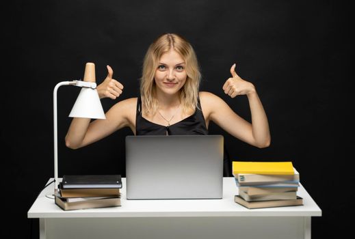 Happy blonde woman sitting at the table with a laptop computer and books and showing gesturing thumb up to a client or collegues over a talk. Office workplace