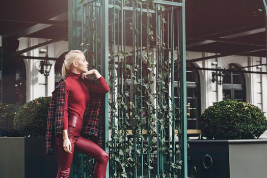 A beautiful, modern, fashionable blonde girl with a red lipstick posing outdoors . Dressed in a red leather leggings, turtleneck and checkered jacket.