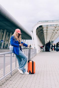 Portrait of a traveler student standing with an orange suitcase near an airport. Young fashionable woman in a blue jeans and jacket, black shirt and white sneakers. Pink baseball cap