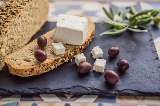 Green and black olives with loaf of fresh bread, feta cheese and young olives branch on olive wood chopping board over dark background.