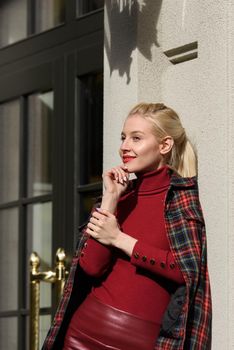 A beautiful, modern, fashionable blonde girl with a red lipstick posing outdoors . Dressed in a red leather leggings, turtleneck and checkered jacket.