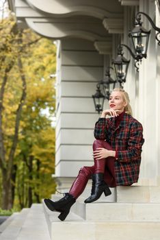fashionable blonde girl with a red lipstick posing outdoors . Dressed in a red leather leggings, turtleneck and checkered jacket. low lace up square toe block heel ankle black leather boots.