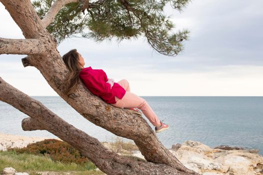 a girl is lying on a tree near the sea shore and looking at a horizon. empty beach, cloudy weather but nature is very beautiful