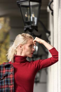 A beautiful, modern, fashionable blonde girl with a red lipstick posing outdoors . Dressed in a red leather leggings, turtleneck and checkered jacket.