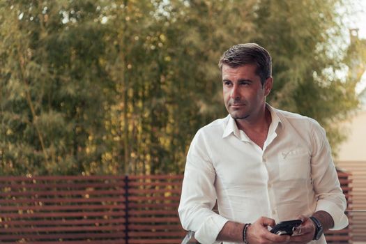 A businessman in casual clothes resting on the balcony of the terrace while using a smartphone. Selective focus. High-quality photo