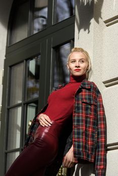 A beautiful, modern, fashionable blonde girl with a red lipstick posing outdoors . Dressed in a red leather leggings, turtleneck and checkered jacket.