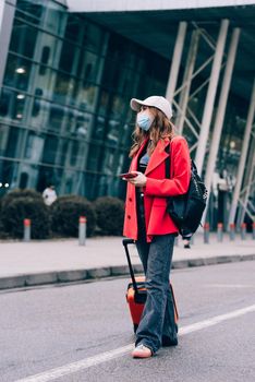 Portrait of a traveler woman in a face mask during virus epidemic walking with an orange suitcase near an airport. Young fashionable woman in a blue jeans and jacket, black shirt and white sneakers