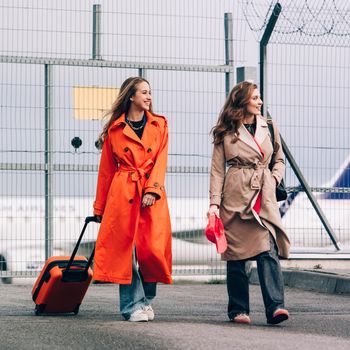 Two happy girls walking near airport, with luggage. Air travel, summer holiday. women dressed in trendy trenches orange and beige. Airport on a background