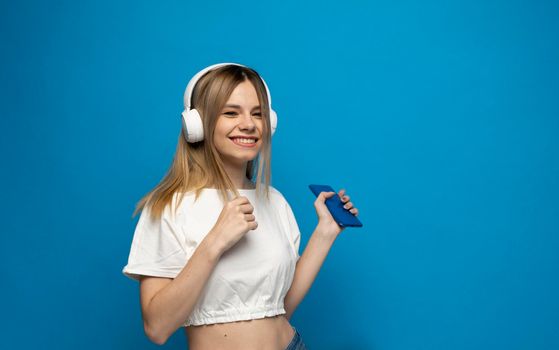 Beautiful young blonde woman with headphones and mobile device listening to music and smiling and dancing, isolated on blue background