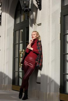 A beautiful, modern, fashionable blonde girl with a red lipstick posing outdoors . Dressed in a red leather leggings, turtleneck and checkered jacket.