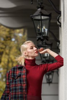 A beautiful, modern, fashionable blonde girl with a red lipstick posing outdoors . Dressed in a red leather leggings, turtleneck and checkered jacket.