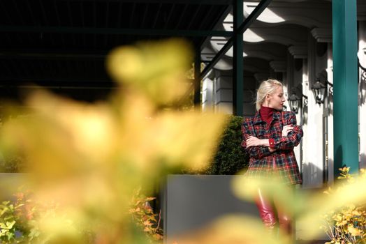 A beautiful, modern, fashionable blonde girl with a red lipstick posing outdoors . Dressed in a red leather leggings, turtleneck and checkered jacket.