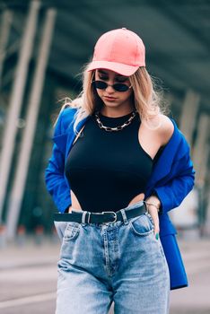Young sexy blonde hipster woman posing on the street. Wearing blue stylish jacket, jeans and baseball hat and sunglasses. Lifestyle portrait