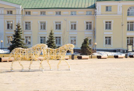 Golden street sculpture in the square of the horse on a frame of light bulbs. Spring, snow melts, in the background is a wall of a multi-storey building with windows. Day, cloudy weather, warm light.