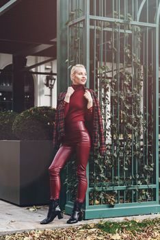 A beautiful, modern, fashionable blonde girl with a red lipstick posing outdoors . Dressed in a red leather leggings, turtleneck and checkered jacket.