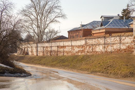 A wet road going down, an old brick wall and roofs of houses on the hill behind. Spring, snow melts, dry grass all around, small snowdrifts and puddles. Day, cloudy weather, soft warm light.