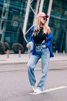 Young sexy blonde hipster woman posing on the street. Wearing blue stylish jacket, jeans and baseball hat and sunglasses. Lifestyle portrait