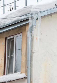 Large ice icicles hang from the drainpipe at the edge of the roof. Against the background of the wall of an old brick house. Large cascades, even beautiful rows. Cloudy winter day, soft light.