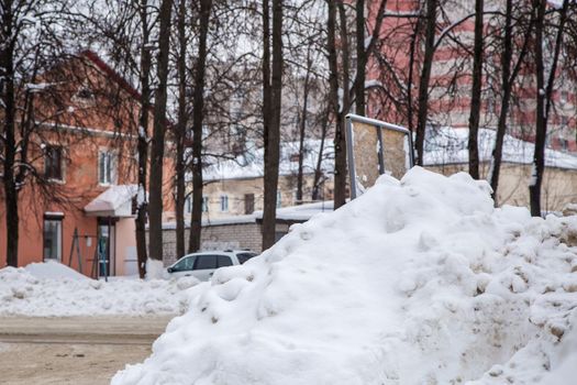 Dirty snowdrift by the road against the backdrop of city houses and trees. On the road lies dirty snow in high heaps. Urban winter landscape. Cloudy winter day, soft light.