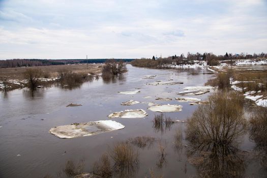 Small white ice floes float down the river slowly. Spring, snow melts, dry grass all around, floods begin and the river overflows. Day, cloudy weather, soft warm light.