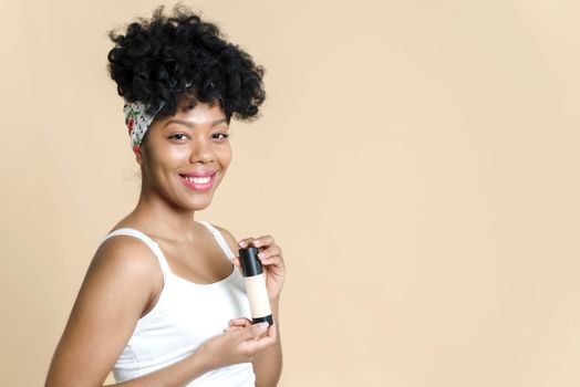 Portrait of a beautiful black woman holding a bottle of liquid foundation on a beige background. African american young woman smiling while holding bottle of foundation.