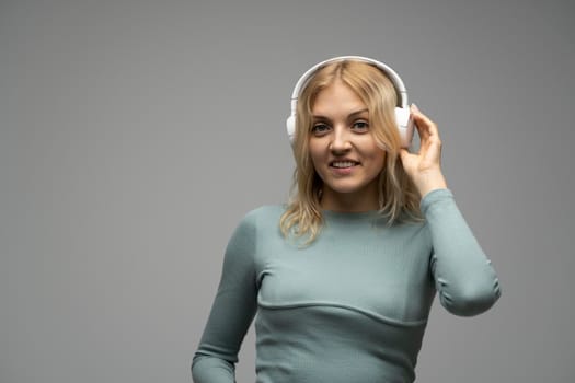 Blonde girl with white headphones listening to music with closed eyes on grey background in studio. She wears grey T-shirt