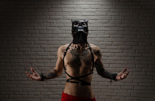 man with a naked torso in a bdsm demon mask with tattoos and leather belts and handcuffs