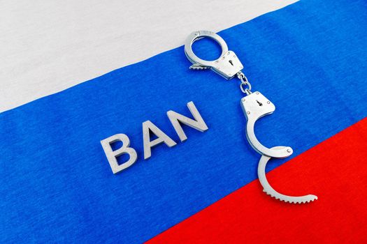 the word ban laid with silver metal letters near opened handcuffs over fabric tricolor flag of the Russian Federation