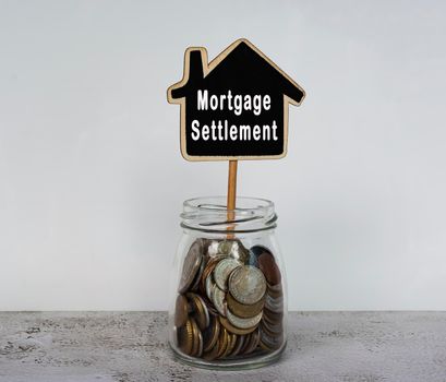 Mortgage settlement text on wooden house model with jar full of coins on a desk.