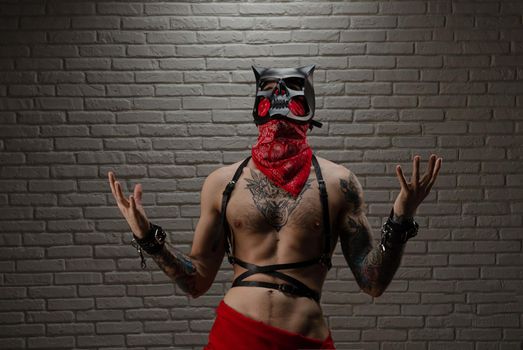 man with a naked torso in a bdsm demon mask with tattoos and leather belts and handcuffs