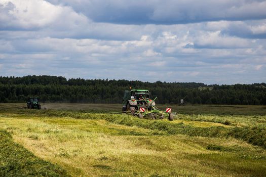 two green haymaking John Deere tractors with Krone plow on summer field before storm - telephoto shot with selective focus in Tula, Russia - July 30, 2019