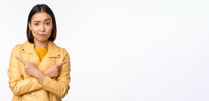 Image of indecisive asian girl, pointing fingers sideways, pointing left and right, choosing variant, deciding, standing over white background. Copy space