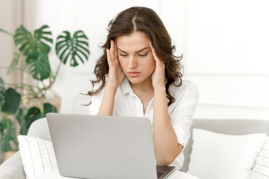 Frustrated sad woman feels tired, headache, worries about a problem while sitting on the sofa with a laptop. Stressed depressed girl worried about reading bad news online or mortgage bills.