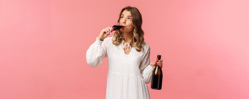 Holidays, spring and party concept. Portrait of young elegant happy blond woman, wear white trendy dress, drinking wine from glass looking up pleased, tasting good drink, hold bottle.