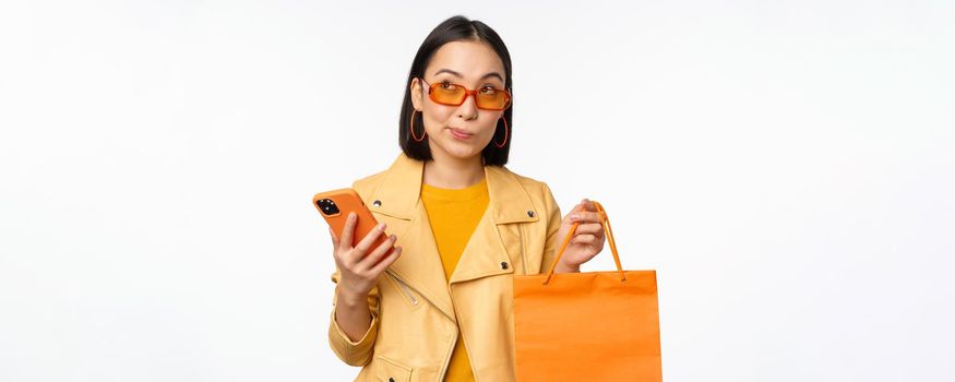 Stylish asian girl shopper, wears sunglasses, holding shopping bag and smartphone, going for discounts in stores, standing over white background.