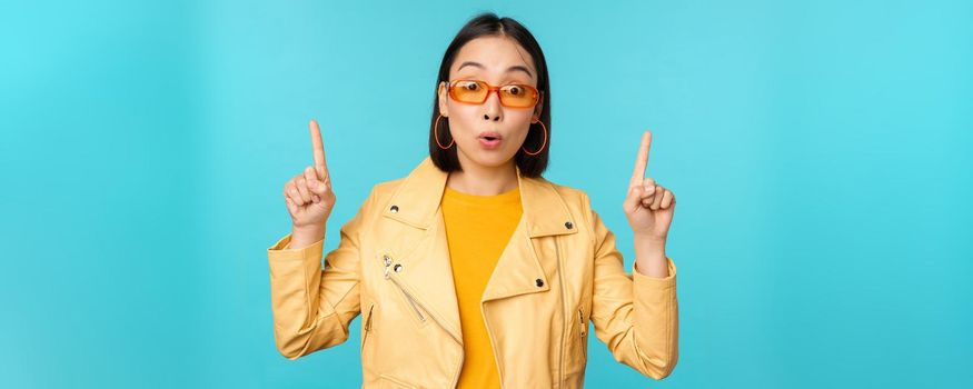 Enthusiastic asian girl in sunglasses, points fingers up, shows banner or logo on top, stands over blue background. Copy space