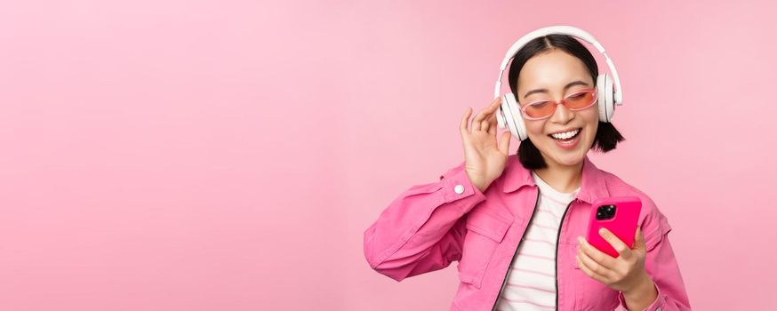 Stylish asian girl dancing with smartphone, listening music in headphones on mobile phone app, smiling and laughing, posing against pink background.