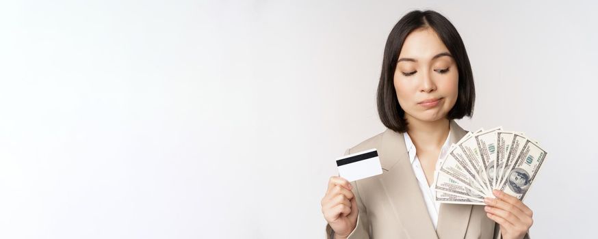 Thoughtful businesswoman, korean corporate woman showing credit card and money cash, dollars in hands, standing over white background and thinking.