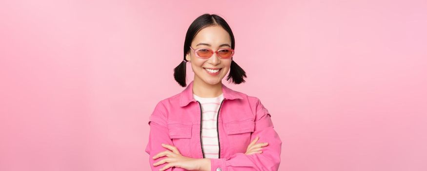 Stylish asian girl in pink clothes and sunglasses, smiling and looking happy at camera, standing over studio background.