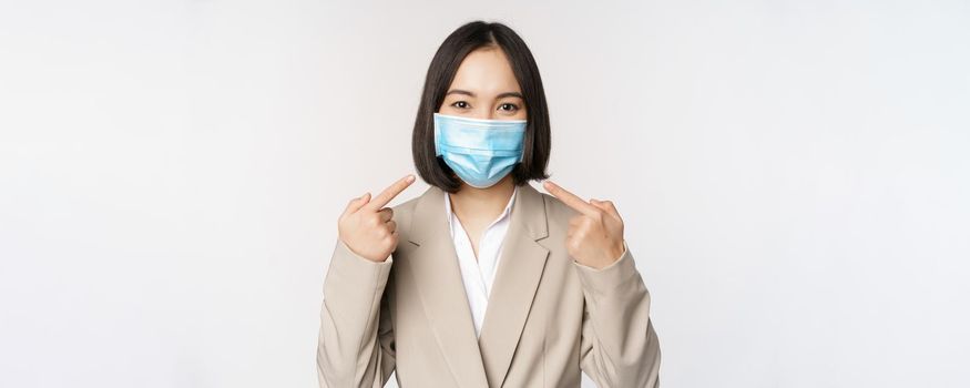 Coronavirus and business people concept. Asian female entrepreneur pointing fingers at medical face mask at workplace, standing over white background.