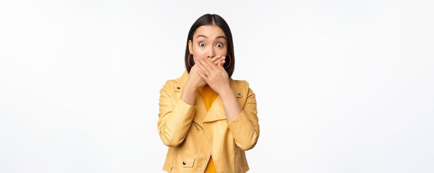 Image of shocked asian girl shuts mouth, close lips and looking speechless, startled face expression, standing over white background.