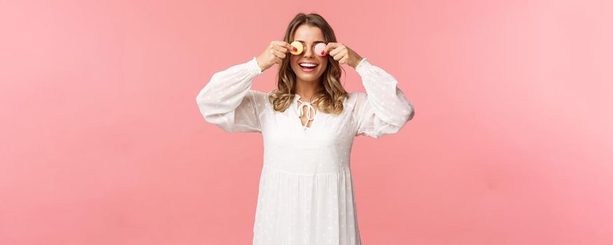 Holidays, spring and party concept. Portrait of carefree funny and cute blond girl in white dress, holding two macarons on eyes and smiling happy, like eating tasty desserts, pink background.