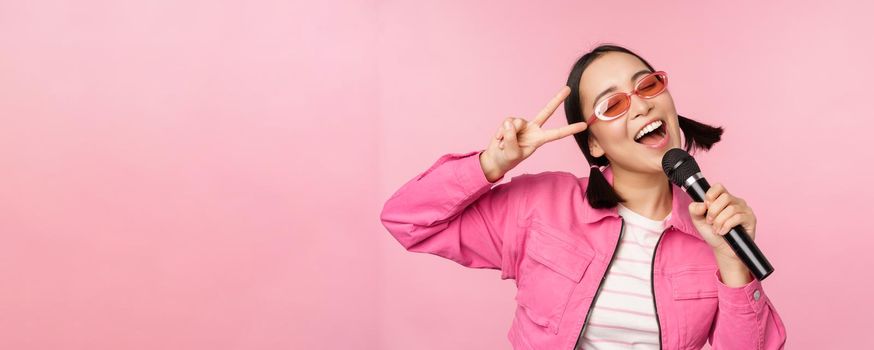 Happy beautiful asian girl singing with mic, using microphone, enjoying karaoke, posing against pink studio background.