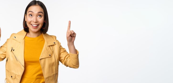 Image of smiling asian brunette woman pointing fingers up, showing advertisement with happy face, posing against white background.