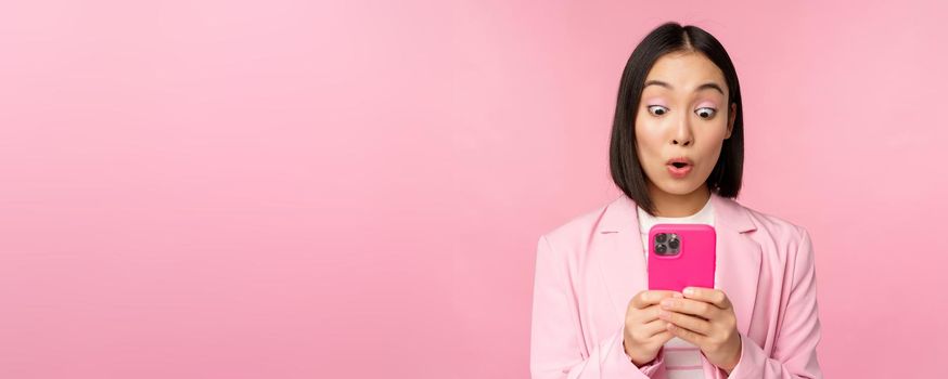 Portrait of asian businesswoman with surprised face, using smartphone app, wearing business suit. Korean girl with mobile phone and excited face expression, pink background.