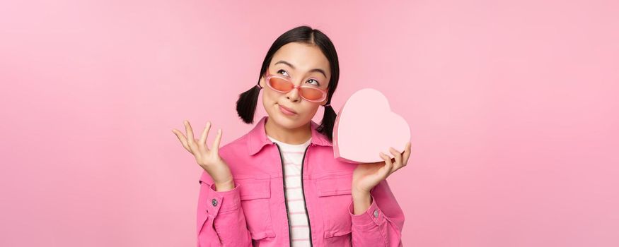 Image of stylish asian girlfriend in sunglasses, guessing whats inside gift box, heart-shaped present, standing over pink background.